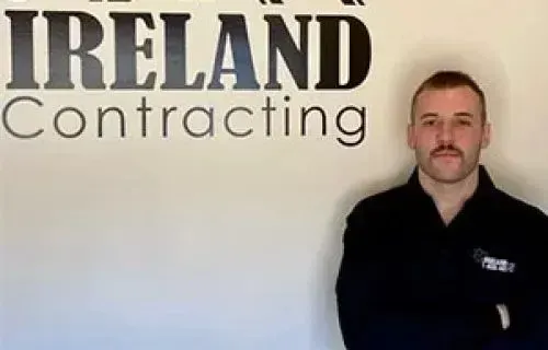 Dan Ireland, electrician at Ireland Contracting, standing in front of the company logo.